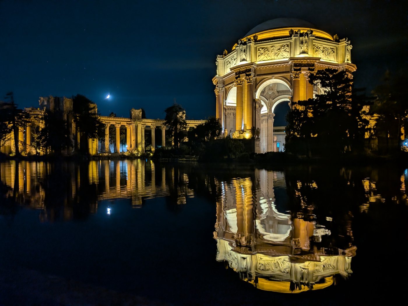 The Palace of Fine Arts lit up at night, reflected in its lagoon under a crescent moon